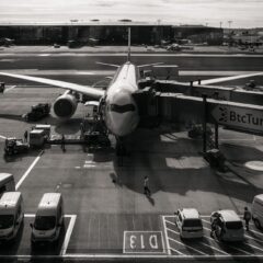 Monochrome image of airplane at airport gate with vehicles and jetway during the day.