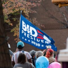 A diverse crowd gathered outdoors holding a Biden 2020 flag during a protest.