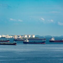 A fleet of cargo ships docked near oil storage tanks along a serene coastline with a clear blue sky above.