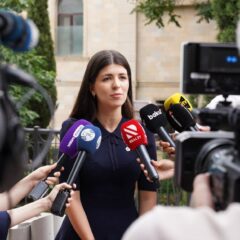 A spokesperson engages with media during an outdoor press interview surrounded by microphones.