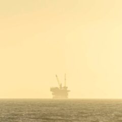 Distant view of an offshore oil platform against a golden sky at sunset over a calm sea.