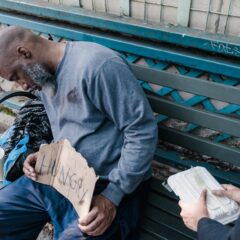 A woman compassionately offers food to a homeless man sleeping on a bench with a 'Hungry' sign.