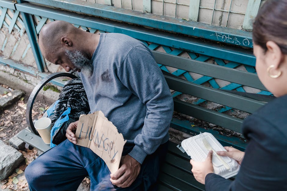 A woman compassionately offers food to a homeless man sleeping on a bench with a 'Hungry' sign.