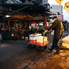 Busy city street market at dusk with a worker pushing a cart of boxes.
