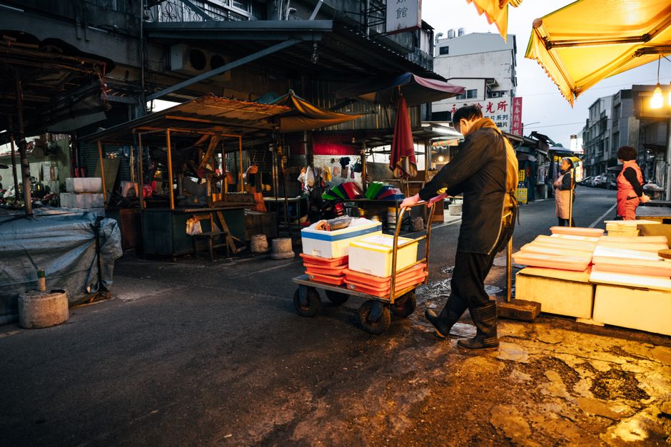 Busy city street market at dusk with a worker pushing a cart of boxes.
