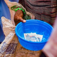 A close-up of a hand holding a blue bowl containing currency notes outdoors.