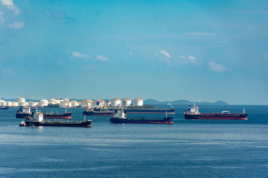 A fleet of cargo ships docked near oil storage tanks along a serene coastline with a clear blue sky above.