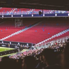 Panoramic view of stadium with empty red seats and crowd gathered for a live match.
