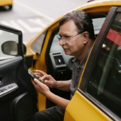Crop Asian man sitting in parked yellow taxi car with open door and using smartphone while looking away