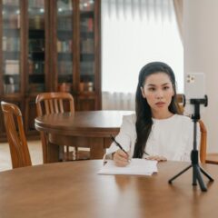 Young Asian woman vlogging in a cozy home office setup using a smartphone and tripod.