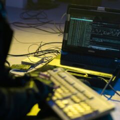 A person typing on a keyboard connected to a laptop showing code, surrounded by wires.