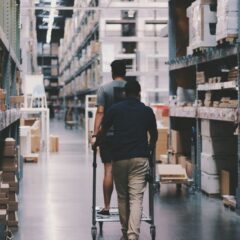Two men maneuver a trolley in a large warehouse filled with boxes and shelves.