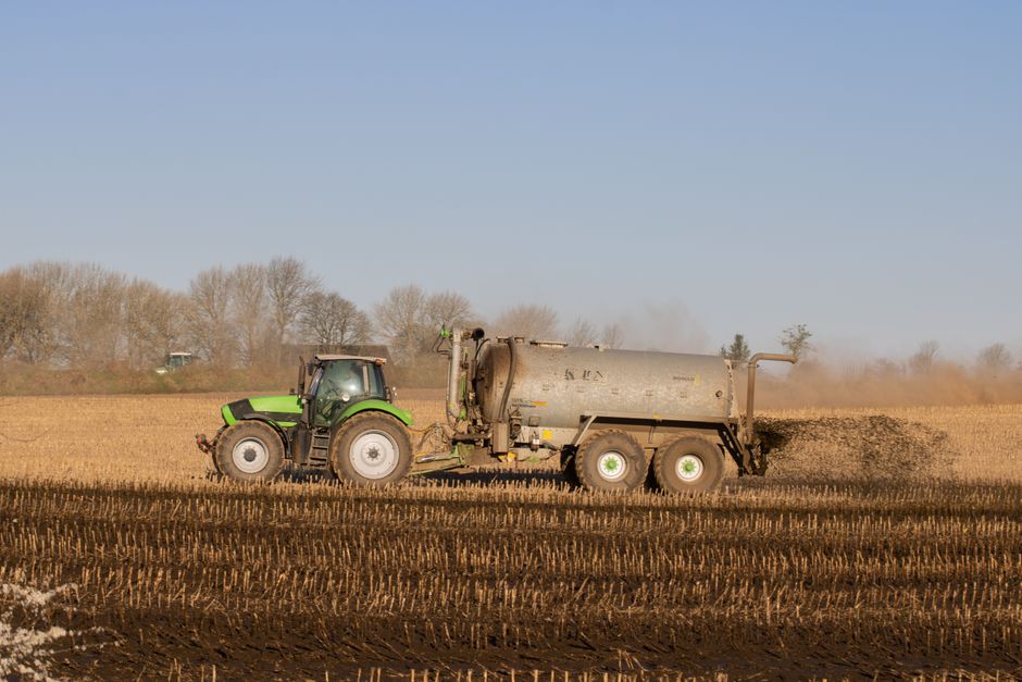 A tractor with a tanker spreading fertilizer over a barren farmland under clear skies.
