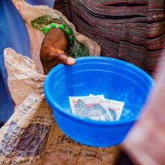 A close-up of a hand holding a blue bowl containing currency notes outdoors.