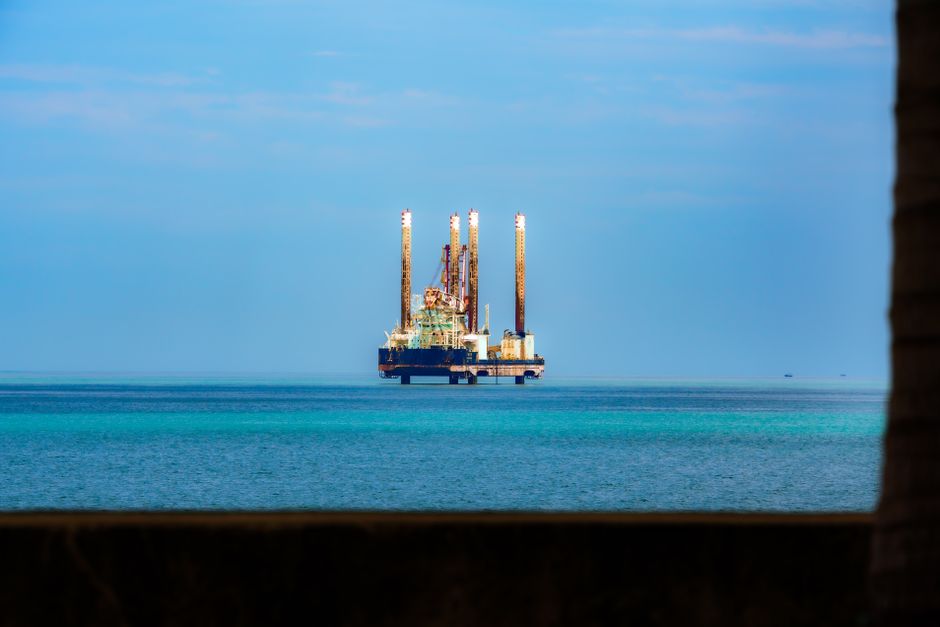 Distant view of an offshore oil rig in a calm, blue ocean under a clear sky.