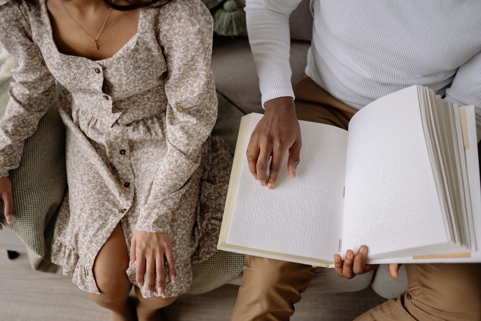 Two people sit together indoors, one reading from a Braille book. Emphasizing accessibility.