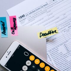 Office desk flat lay showing tax documents, calculator app on smartphone, sticky notes, and paperclips.