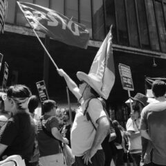 Crowd holds flags and signs during protest in urban setting.