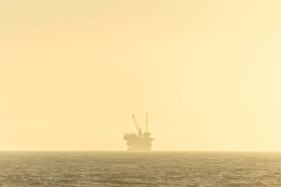 Distant view of an offshore oil platform against a golden sky at sunset over a calm sea.