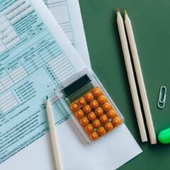 Flat lay of tax forms, calculator, pencils, and clips on green surface, ideal for finance or accounting themes.