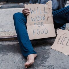A homeless person sitting on the street holding a cardboard sign with the message 'Will work for food'.